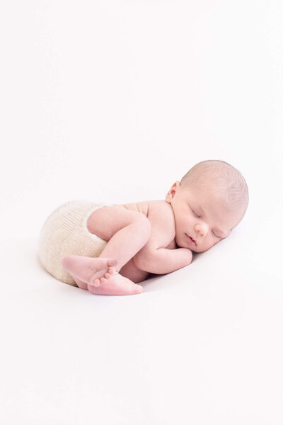 A baby wearing a delicate, frilly dress and a floral headband lies on a white surface. The baby is smiling softly, with their head turned to the side. The setting is simple and minimalist.