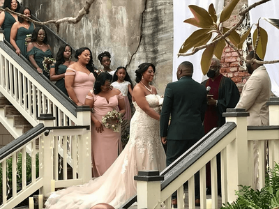 A bride and groom stand facing each other during their wedding ceremony on an outdoor staircase, surrounded by bridesmaids in pink dresses and groomsmen, with an officiant and lush greenery nearby.