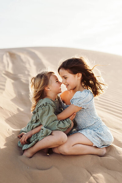 Two little girls are sitting on sand dunes, hugging each other and smiling."