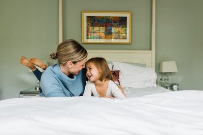 A woman and a young girl smiling at each other while lying on a bed in a room with neutral colors during a family photography session in Pittsburgh.