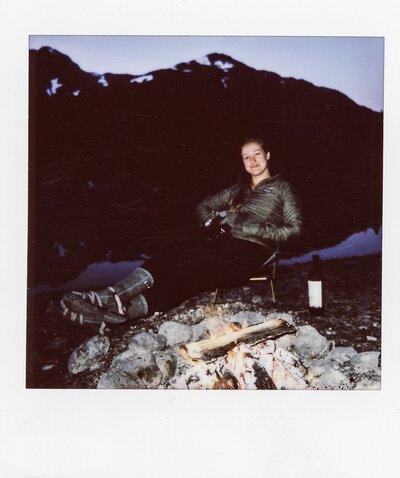 Polaroid of JayneMayAgnes sitting next to a campfire with a bottle of wine