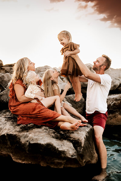 Family Photographer, a father lifts his daughter in the air at beach, mom and sisters sit close on the rocks