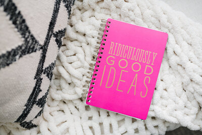 Pink notebook with the title 'ridiculously good ideas' laying on top of a white blanket