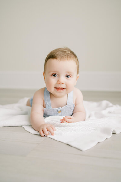 Baby boy with blue eyes lays on his belly over a white blanket and smiles at the camera during a studio photoshoot.
