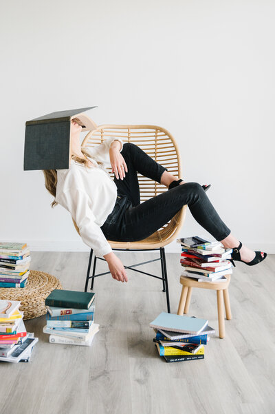 Sarah Klongerbo leaning back in a chair surrounded by stacks of books with an open book laying on top of her face