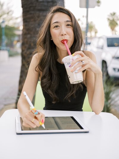 Woman with red lipstick looking at the camera and drinking boba at an outdoor table while working on her ipad