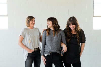 Three women casually dressed, laughing and leaning on each other in a minimal studio space at Photobooth.