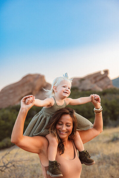 Mom has young daughter in a green dress on her shoulders, in front of red rock formations.