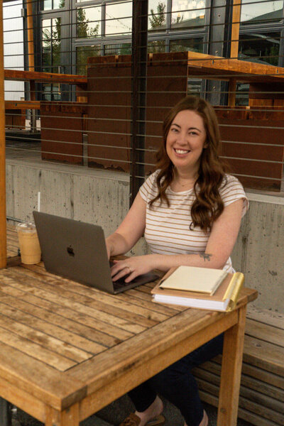 Virtually Robyn, a woman and virtual assistant, with long brown hair, wearing a striped shirt, sits at an outdoor wooden table with a laptop, notebooks, and an iced drink, smiling at the camera.