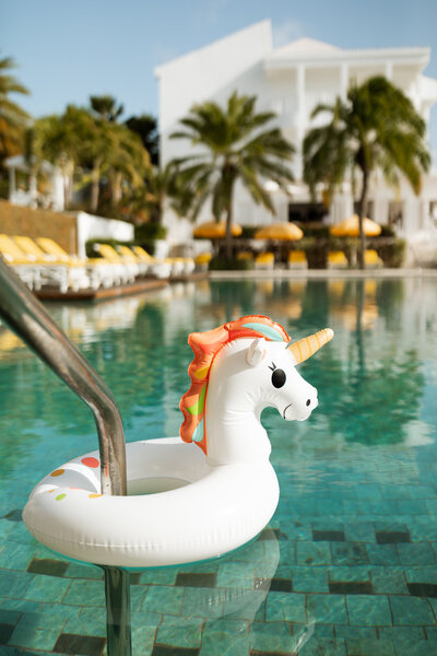 A unicorn pool float in a hotel pool, framed by palm trees and yellow lounge chairs