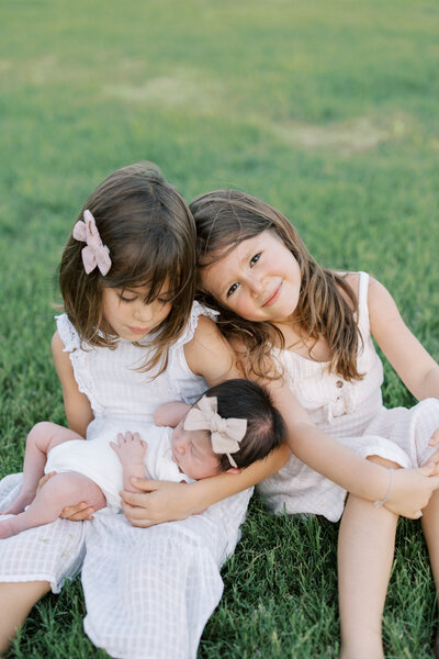 Two older sisters snuggle their new baby sister while one looks at the baby and the other at the camera