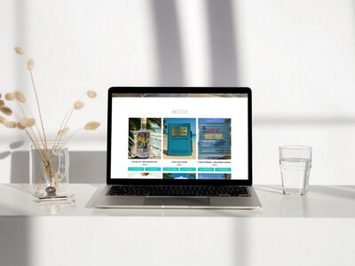 Laptop on a white desk displaying an eCommerce website with rum-themed products, including a rum bottle, rum lovers journal, and a Caribbean cookbook, lit by natural sunlight with a glass of water and dried florals beside it.