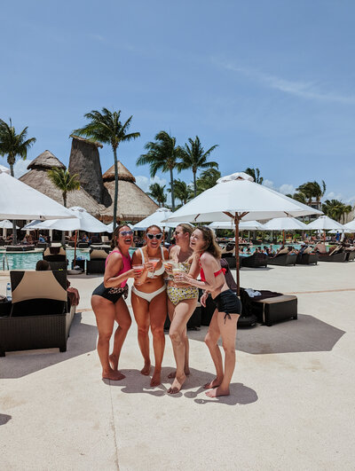 Four women in swimsuits stand by the pool under large white umbrellas, holding drinks and laughing together in a tropical resort setting. Palm trees and lounge chairs surround them, providing a relaxed vacation atmosphere.