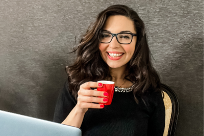 An image of Rania Fayad wearing glasses and holding a red coffee cup smiling at the camera. She is wearing a black jumper with pearls around the neckline, red nail polish and her hair is dark brown, wavy and down.