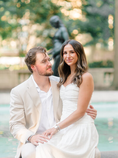 A stylish couple poses beside a tranquil fountain in Rittenhouse Square, Philadelphia. The woman, dressed in a white ruched dress, sits gracefully on her partner’s lap while he gazes at her lovingly. The bronze sculpture and soft greenery in the background add an elegant, timeless ambiance to this romantic portrait by Elizabeth Kane Photography, a Luxury Philadelphia Wedding Photographer.