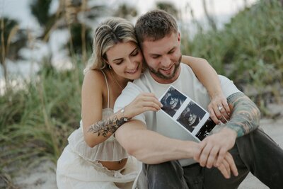 couple smiles and holds ultrasound picture on the beach in south florida