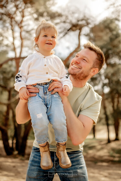 Papa tilt zijn dochter de lucht in en kijkt stralend naar haar bij Herperduin in het bos
