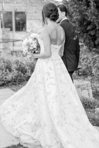 Black-and-white image of a bride in a stunning floral lace gown walking hand-in-hand with her groom in Dripping Springs, Texas. The serene moment captures the beauty and elegance of the couple on their wedding day. Felisha, a New England-based destination wedding photographer, specializes in capturing timeless moments in Texas, New England, and worldwide locations.