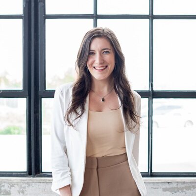 Bri Carlesimo smiling in a white blazer and beige top, standing in front of large industrial-style windows.