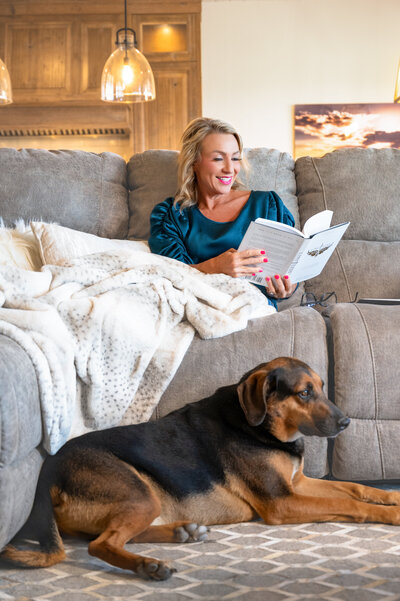Professional real estate agent seated in a designer lounge, book in hand, surrounded by chic, modern decor, dog, and natural light.