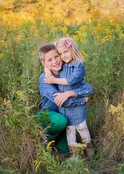 older brother and younger sister both wearing denim shirts hug in a field of tall grass and yellow flowers