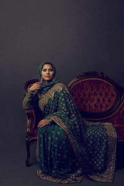 Fine art portrait of a woman in a green and gold sari sitting on an antique sofa. Houston, TX.