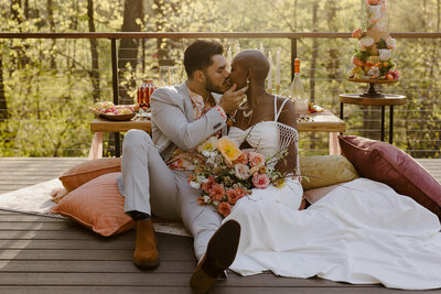 A couple kisses while sitting on a porch, greenery behind them.