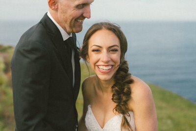 bride and groom laugh beside a sea cliff