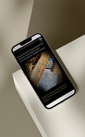 A smartphone displaying an e-commerce mockup for Cuttlebrook Knives, featuring an image of a handcrafted knife with intricate patterns on the blade. The text on the screen reads, "Exceptional Handcrafted Knives for Chefs and Enthusiastic Home Cooks," highlighting the artisanal quality of the product. The phone is placed on a light-colored surface, adding to the sleek and professional presentation.