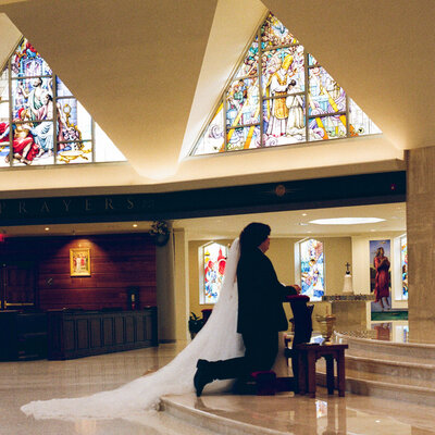 Bride and groom kneeling during a Catholic wedding ceremony with stained glass windows in the background, captured in a destination wedding documentary style by Emily Ambie.
