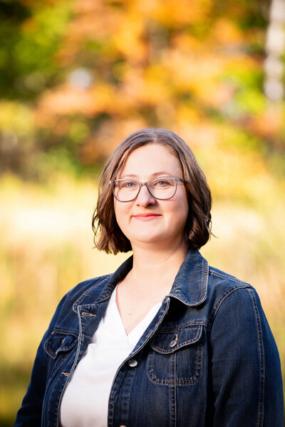 Close-up of Kasey Pierce, wearing a white blouse, denim jacket, and glasses, in front of a background of autumn trees