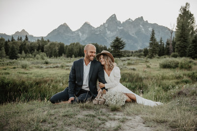 boho bride in a casual white dress and boho hat sits next to her groom  who is wearing a blue suit in a field in front of the Teton mountain range