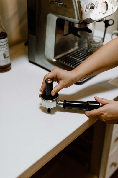 A person tamping ground coffee into a portafilter next to an espresso machine on a kitchen counter—fueling up before diving into their white label design course.