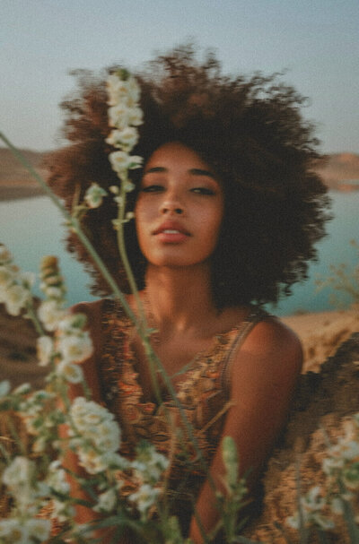 A woman with curly hair sits outdoors near a body of water, surrounded by white wildflowers and hills—a serene moment that captures the inspiration behind Sweet Pea Ink Creative’s brand design.