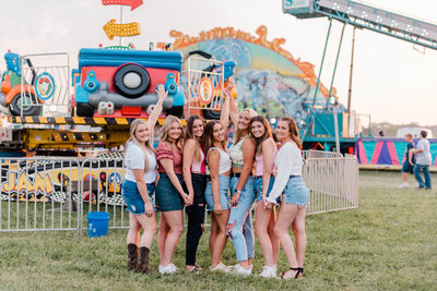 Rachel B Photography's Senior Rep Team poses together for the camera with their arms in the air in front of a carnival ride.