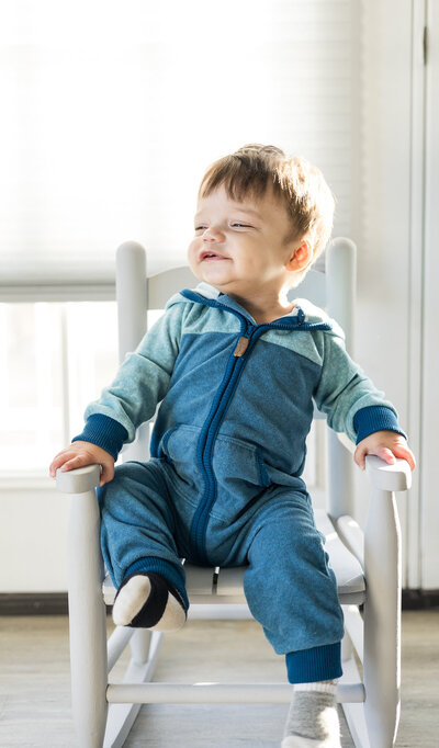 12 mo old boy with short dark brown hair, sitting in a light gray rocking chair, looking away from the camera smiling, and wearing a blue zip one piece.