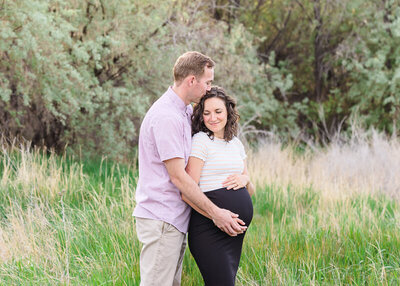 A husband wearing a pink button down shirt and khaki pants holds his pregnant wife wearing a white striped shirt and black skirt as they stand in the tall grass at North Lake Park in Lehi. Captured by Utah maternity photographer Melissa woodruff photography