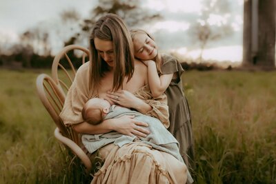 Woman seated on a chair in a field, breastfeeding a baby wrapped in a blue blanket. A young girl in a dress stands beside, eyes closed, leaning her head on the woman's shoulder.