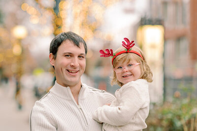 Young daughter in a white dress and red reindeer antlers kissing Dad's cheek during their Christmas photos in Chicago, IL