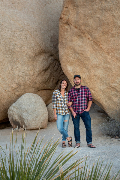 Becky & Brian, elopement photographers, stand in front of a boulder in Joshua Tree, smiling at the camera.