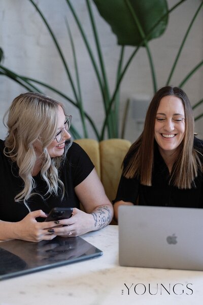 Jazmin and Alex are sitting in a curved booth. Both are handling client business—Jazmin on her phone, and Alex on her laptop showing Jazmin something. They're smile at each other.