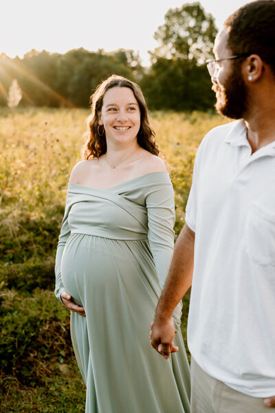 Pregnant mom holding the hand of her husband and smiling at her while walking in a golden lit field in Annapolis Maryland.