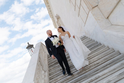 Bride and groom share joyful moment on white marble stairs against blue sky.