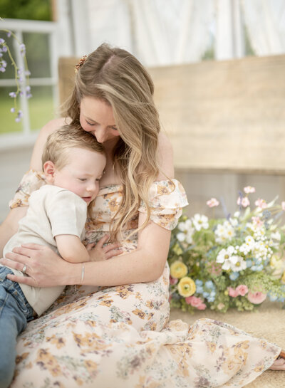Mother holding her toddler close while sitting near pastel floral arrangements during a spring family session with a Kansas City family photographer.