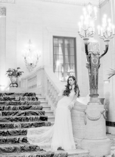 Black and white photograph of a model in a white all lace wedding gown with long lace sleeves that open up and hang at the wrists and a long lace train which is resting on the grand staircase the model is on. Model has long waist length dark brown vintage waved hair with long white feathered Oscar de la Renta earrings. Model is standing leaning against the stone railing of the 1800's staircase with grand candelabras that the bottom of the staircase and at the top. Photographed by wedding photographers in Charleston Amy Mulder Photography