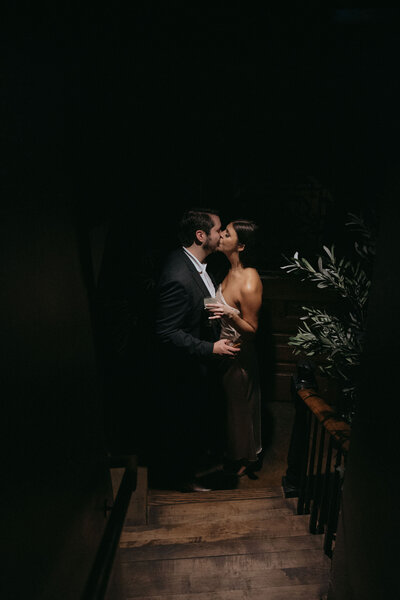 A couple shares a kiss at the bottom of a dark stairwell. They are holding cocktails in their hands, about to enter a speakeasy for their engagement photo session at Smoked in South Carolina.