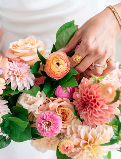 A woman’s hand with gold rings gently holds a bouquet of vibrant flowers, including pink and peach dahlias, roses, and ranunculus, against a white background. The arrangement is lush with green leaves, conveying a sense of elegance and freshness.