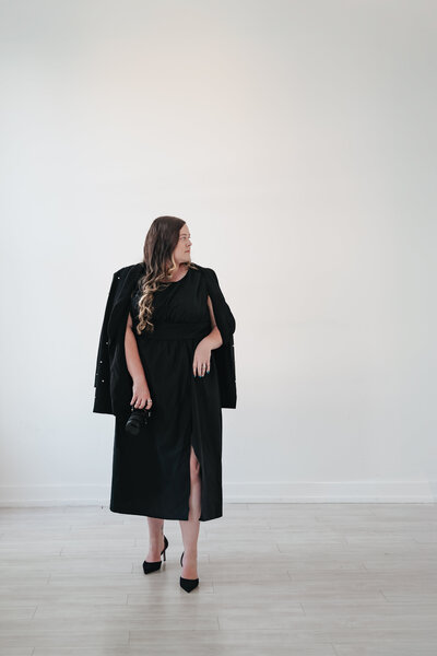 Portrait of the owner of Focused On Life Photography wearing a pearl-embellished black jacket and long black dress, confidently holding a camera with modern pampas grass decor, capturing a bold and stylish fashion-forward branding aesthetic.