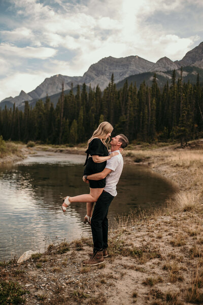 Man lifts woman up in front of a lake and mountains