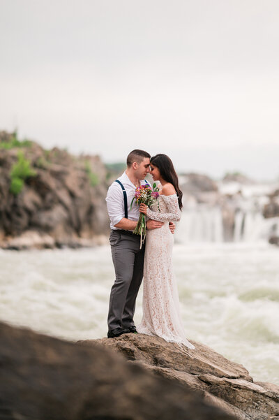 A newlywed couple poses for a stunning portrait at the water's edge during a session at Great Falls National Park in Virginia, captured after hiking to the base of the falls by Nathan Sottung Photography